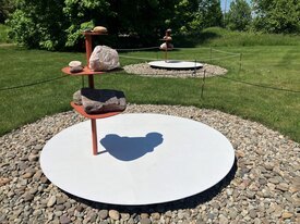 A sculpture of a large white disk, with red poles jutting out that has rocks arranged on platforms. On the white disk is a shadow of Buddha.
