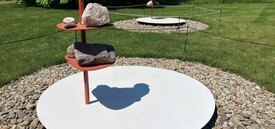 A sculpture of a large white disk, with red poles jutting out that has rocks arranged on platforms. On the white disk is a shadow of Buddha.
